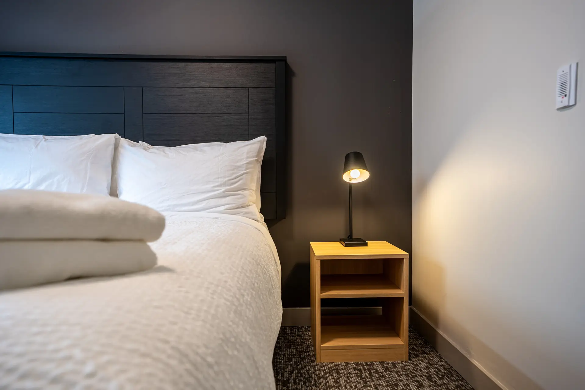 Close-up of a white bed with stacked towels and a bedside lamp in a hotel room.
