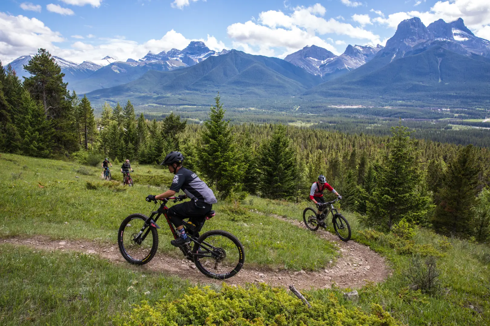 Mountain bikers descend a scenic trail in Canmore, Alberta, with the Three Sisters mountains in the background.