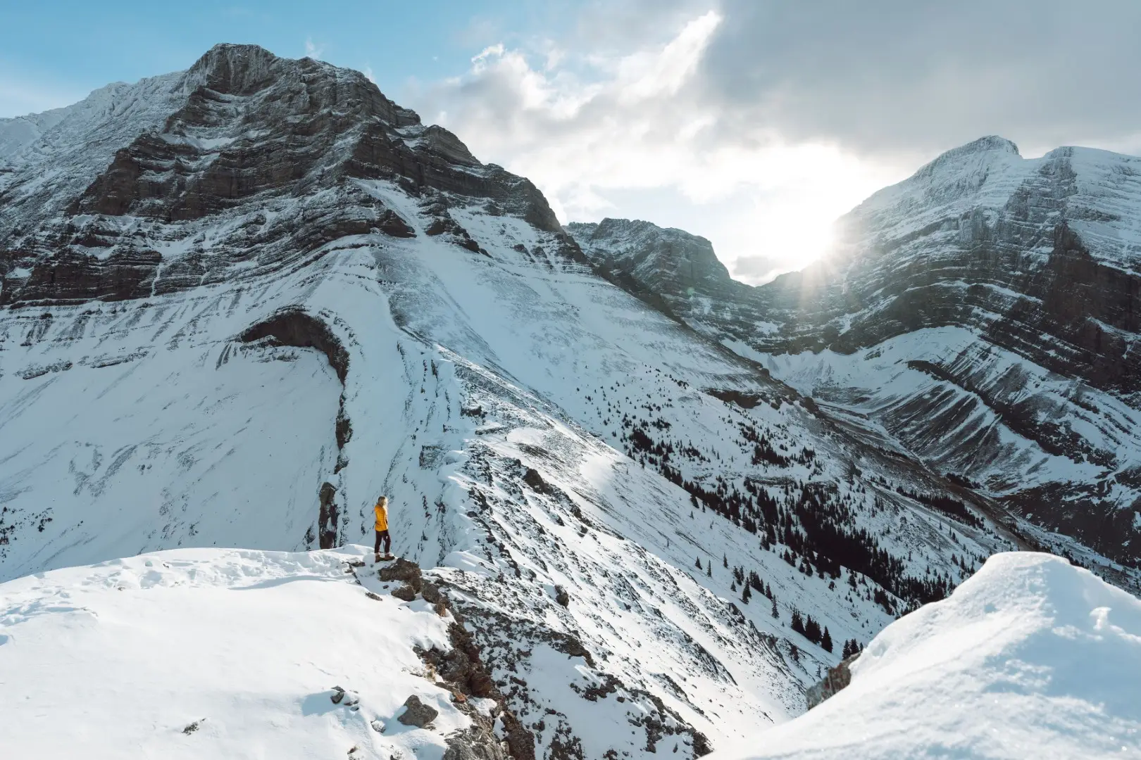 Person in yellow jacket on snowy mountain ridge near Canmore, Alberta, with sunbeams.