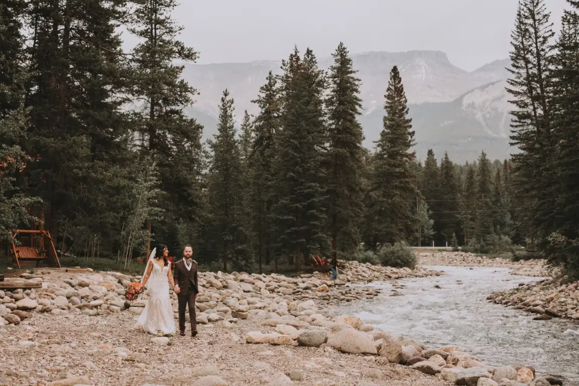 Bride and groom walking along rocky riverbank with forest and mountains behind.