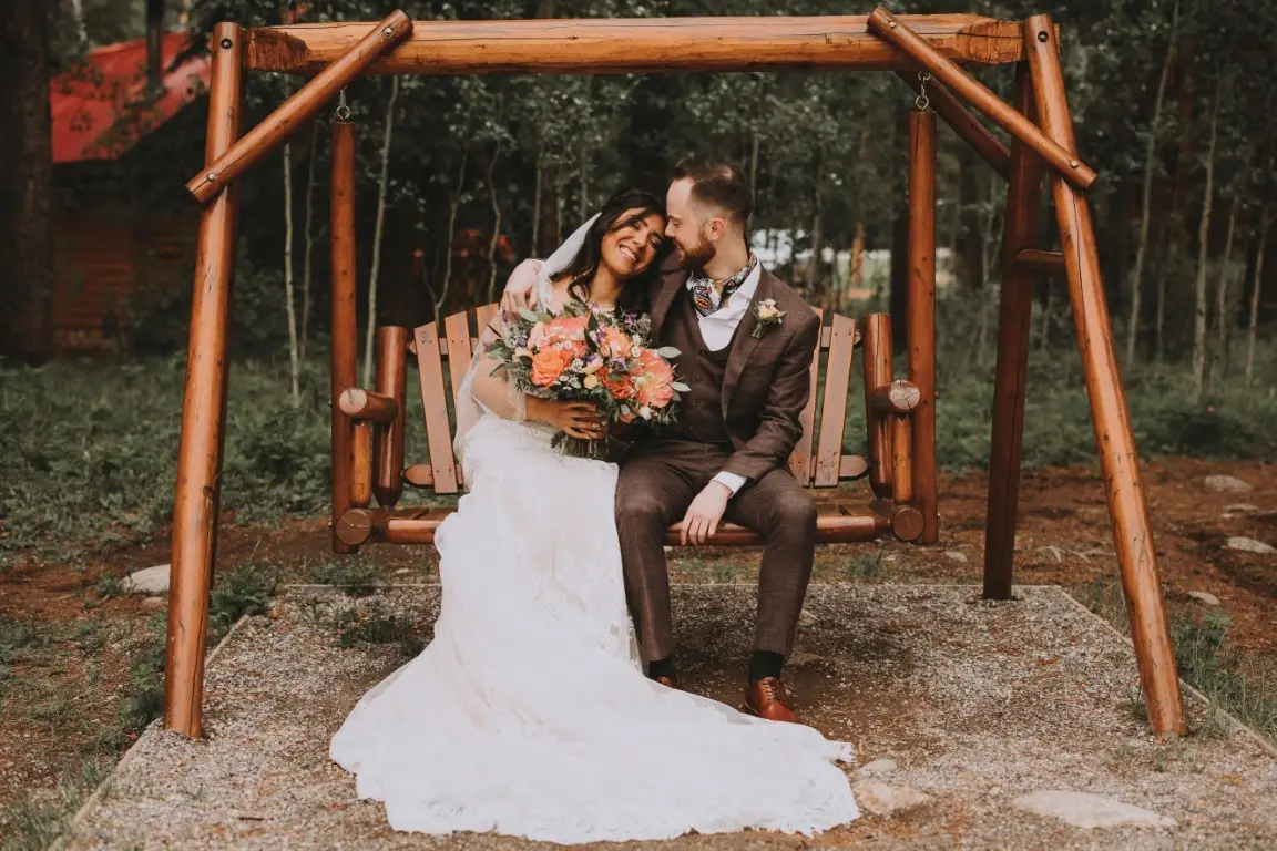 Bride and groom sitting on wooden swing, smiling, holding a bouquet.