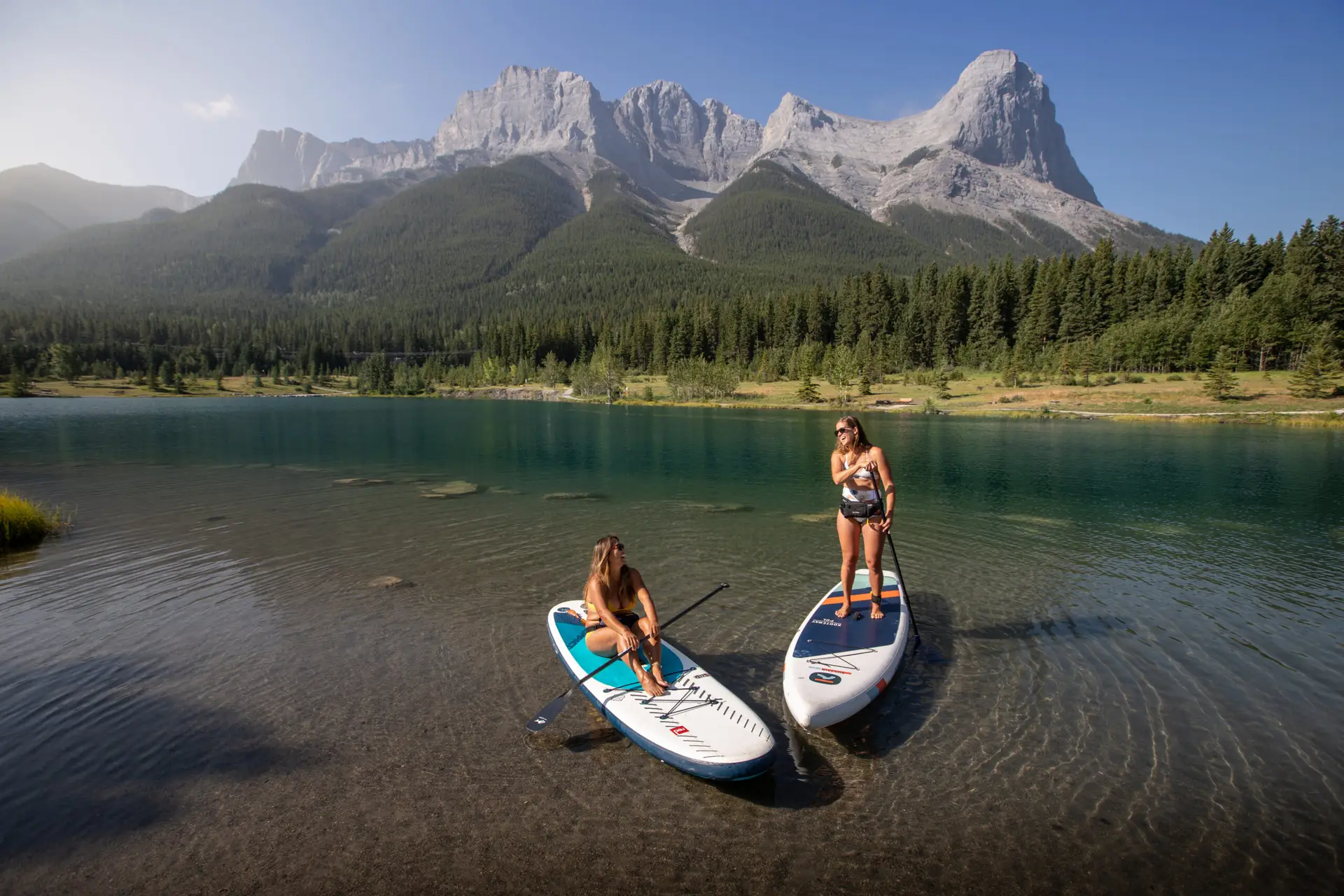 Two women paddleboarding on a clear lake with mountains in Canmore, Alberta.