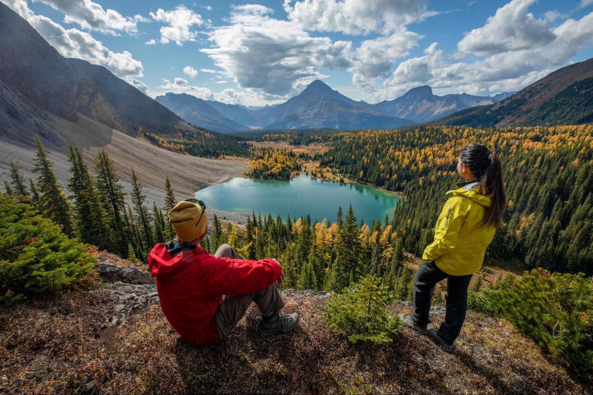 Two hikers admire a turquoise lake surrounded by golden larches during a Canadian Rockies larch hike.