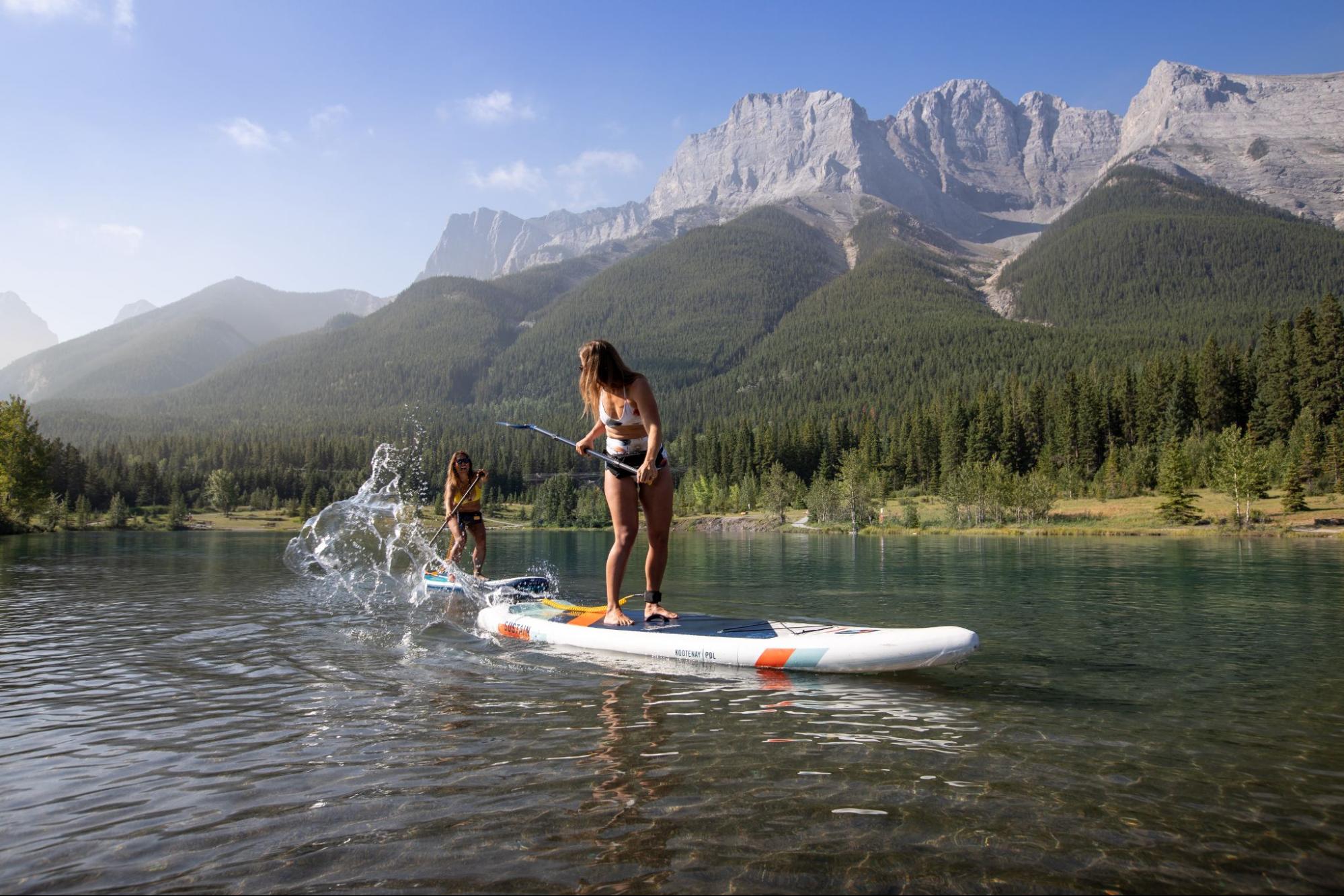 Two women paddleboarding on a clear lake with mountains in the background near Banff.