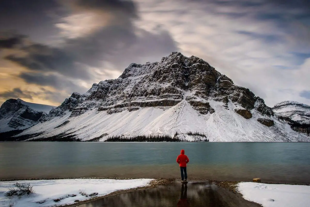Person in red jacket by Banff lake with snow-capped mountain, Banff vs Canmore winter