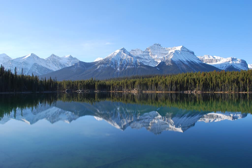 Snow-capped mountains reflecting in a serene lake surrounded by pine trees, a beautiful Banff and Canmore lake.