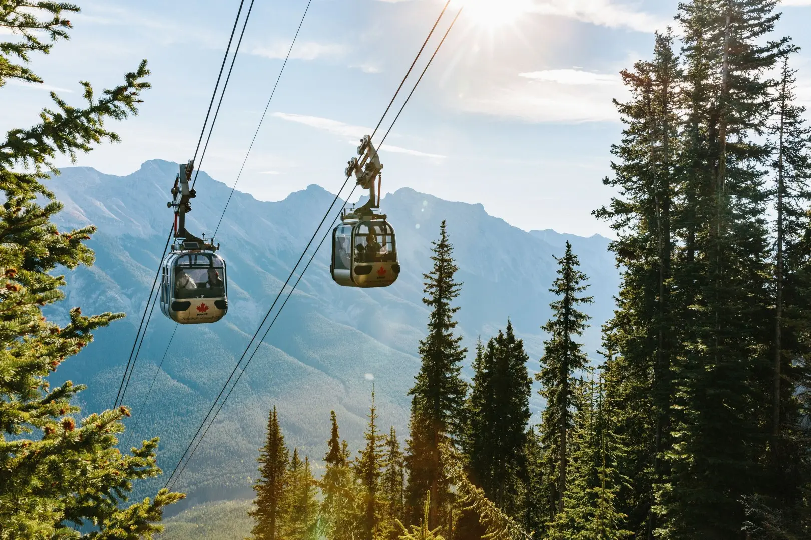Two gondolas suspended over forested mountains under a bright, sunny sky.